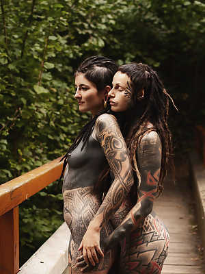 Heavily tattooed lesbians hold each other while totally naked on a bridge