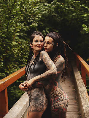 Heavily tattooed lesbians hold each other while totally naked on a bridge