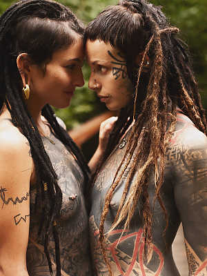 Heavily tattooed lesbians hold each other while totally naked on a bridge