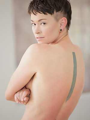 Short haired dyke Jiz Lee strips for nude poses against a cinder block wall