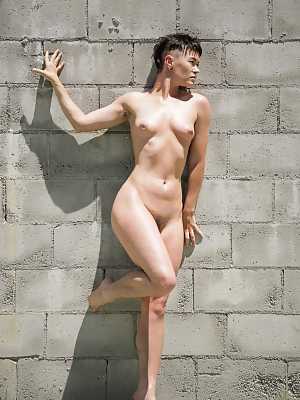 Short haired dyke Jiz Lee strips for nude poses against a cinder block wall